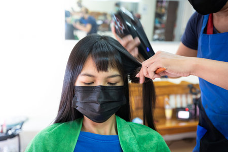 Young asian woman wearing protection mask getting a drying hair with hair dryer by hairdresser at hair salon. Hairdresser drying hair to client. Beauty salon, hair care concept.の写真素材