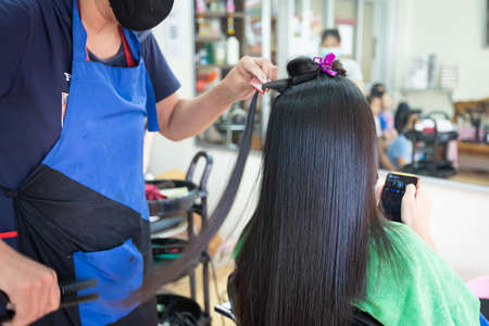 Young asian woman using smartphone while she getting a straight hair with hair straightener by hairdresser. Beauty salon, hair care concept.の写真素材