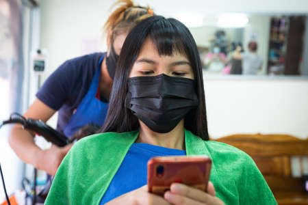 Young asian woman wearing protection mask and using smartphone while she getting a drying hair with hair dryer by hairdresser. Hairdresser drying hair to client at hair salon.の写真素材