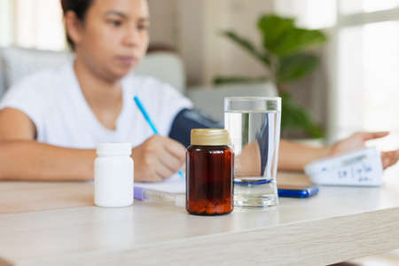 Close up on pills bottle and a glass of water while asian young woman is checking blood pressure and heart rate with digital pressure gauge by herself at home. Health and Medical concept.の写真素材