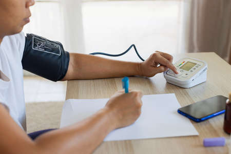 Asian young woman is checking blood pressure and heart rate with digital pressure gauge by herself and record on paper at home. Health and Medical concept.の写真素材