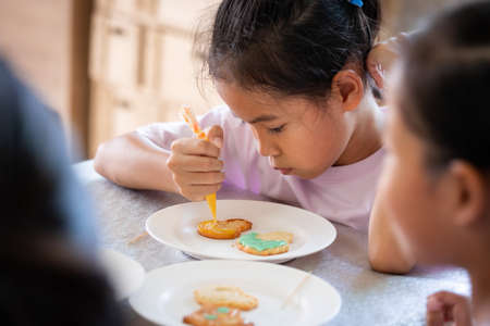 Close up asian child girl squeezing icing cream to decorate cookies with friend with fun. DIY homemade kid art and craft concept.の写真素材