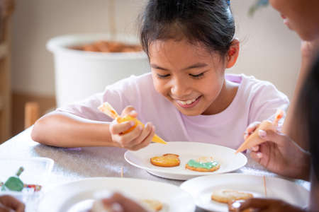 Close up asian child girl squeezing icing cream to decorate cookies with friend with fun. DIY homemade kid art and craft concept.の写真素材
