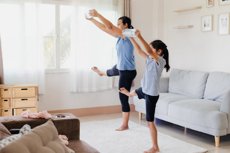 Asian young mother and her daughter doing stretching fitness exercise yoga and using the bottle of water to be dumbbells together at home in daily routine.の写真素材