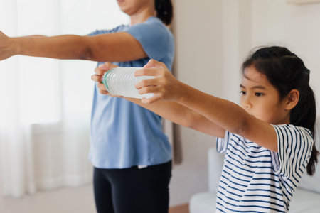 Asian young mother and her daughter doing stretching fitness exercise yoga and using the bottle of water to be dumbbells together at home in daily routine.の写真素材