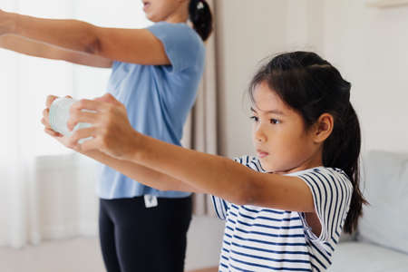 Asian young mother and her daughter doing stretching fitness exercise yoga and using the bottle of water to be dumbbells together at home in daily routine.の写真素材