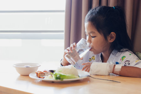 Sick asian child girl drinking a glass of water after ate healthy food for lunch by herself while stay in private patient rooms in the hospital. Healthcare and lifestyle concept.の写真素材