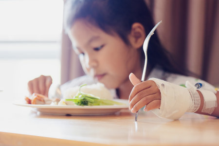 Sick asian child girl eating healthy food for lunch by herself while stay in private patient rooms in the hospital. Healthcare and lifestyle concept.の写真素材