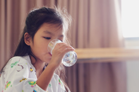Sick asian child girl drinking a glass of water after taking medicine while stay in private patient rooms in the hospital. Healthcare and lifestyle concept.の写真素材