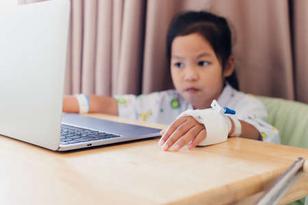 Close up on sick child's hand who have IV solution while child studying online video call with laptop computer in the hospital. Isolation homeschooling and distance learning.の写真素材