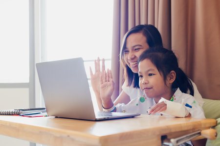 Asian mother and sick daughter wave their hands say hello to family in video call online on computer laptop while stay in hospital. Social distancing during pandemic.の写真素材