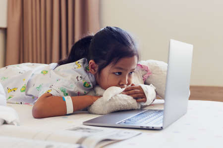 Sick asian child feel bored and lying on bed while learning online class with teacher and friend when stay in private patient rooms in the hospital. Isolation homeschooling and distance learning.の写真素材