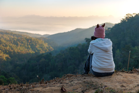Rear view of asian woman sitting on the ground looking on the view of beautiful nature on the top of mountain. People enjoying with panoramic view in the morning time.の写真素材