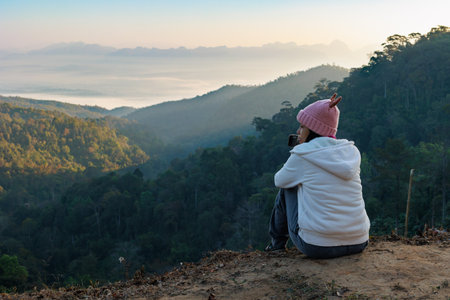 Rear view of asian woman sitting on the ground looking on the view of beautiful nature on the top of mountain. People enjoying with panoramic view in the morning time.の写真素材