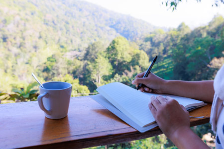 Asian woman sitting on balcony and writing idea on notebooks near fresh nature with hot drink in the morning time.の写真素材
