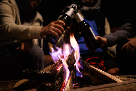 Group of asian friends sitting around campfire camping near bonfire enjoying and drinking beer and relaxing on winter vacation at the night time.の写真素材