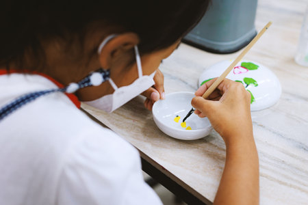 Asian child girl using paint brush is painting on ceramic bowl with oil color. Kids arts and crafts creative activity class in workshop. DIY for kids, home activities, handmade decorations.の写真素材