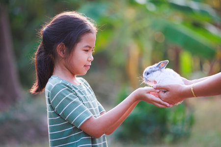 Asian child girl getting adorable bunny from her mother hand and she  playing and caring cute little rabbit holland lop in the garden. Symbol of Easter day.の写真素材