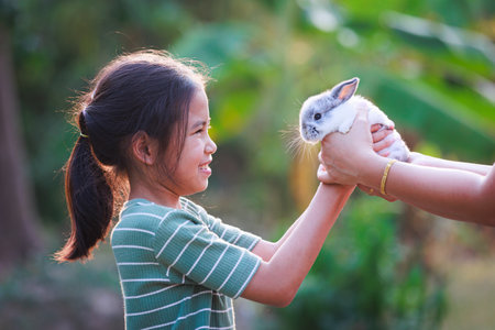 Asian child girl getting adorable bunny from her mother hand and she  playing and caring cute little rabbit holland lop in the garden. Symbol of Easter day.