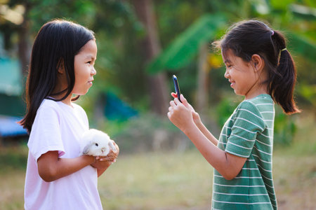 Asian child girl holding a adorable bunny Holland lop on hand and her friend using smartphone taking a picture. They playing with pet together with fun in the yard.の写真素材