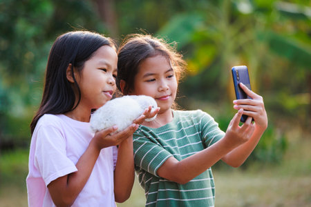 Two asian girls taking a picture and playing with adorable bunny. Sister siblings holding a little rabbit Holland lop on hand and using smartphone selfie together with fun in the garden.の写真素材