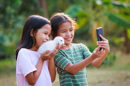 Two asian girls taking a picture and playing with adorable bunny. Sister siblings holding a little rabbit Holland lop on hand and using smartphone selfie together with fun in the garden.の写真素材