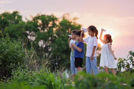 Cute asian little girl and her siblings playing with paper airplane toy together with fun. Group of asian kid throwing paper plane and playing together in the countryside at the sunset time.の写真素材