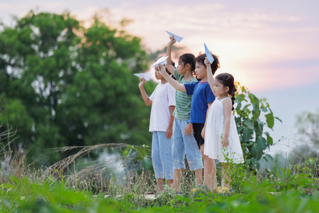 Cute asian little girl and her siblings playing with paper airplane toy together with fun. Group of asian kid throwing paper plane and playing together in the countryside at the sunset time.の写真素材