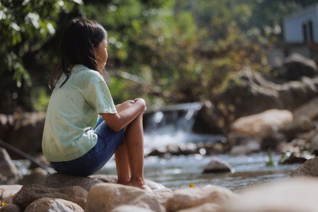 Sad asian child girl sitting on the rock beside the river alone. Children feeling upset while traveling with family.の写真素材