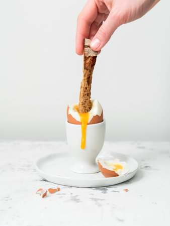 Female hand putting crouton in yolk of soft-boiled egg in eggcup on white table and with white...