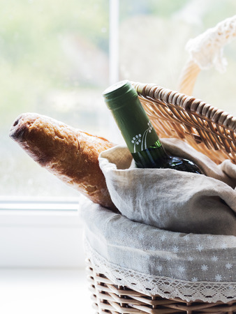 Baguette and bottle of wine in vintage wicker basket on window sil sill with green plantsの写真素材