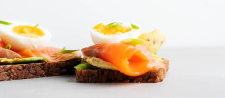 Healthy open sandwiches on multigrain wholegrain  toast with avocado, salmon, eggs, herbs, sunflower seeds over white plate on concrete background with copy space. Healthy protein food. Wide banner.の写真素材