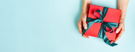 Female hands is holding present red box with green bow on pastel green background with copy space....