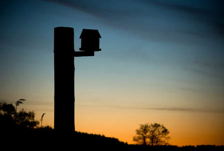 Silhouette of a bird house on a pole against orange and blue sky in sunsetの写真素材
