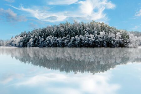 small hill with conifers reflected in water on bright winter dayの写真素材