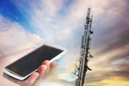 Hand holding white mobile phone aiming at telecommunication tower on blue sky in background