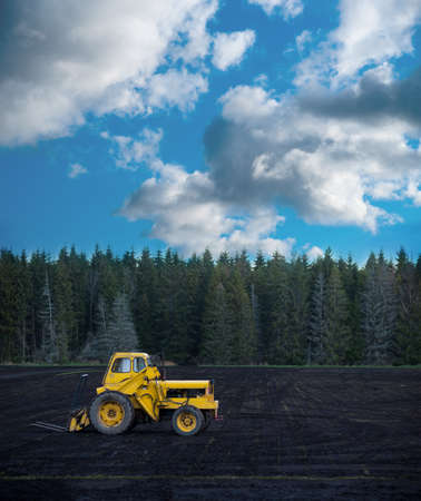 Vintage yellow tractor on dark field with soil, blue sky with clouds and forest of conifers in backgroundの写真素材