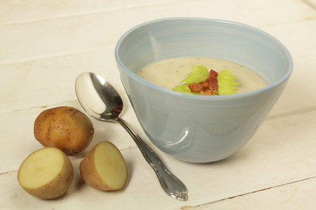 Potato soup in a blue bowl with fresh potatoes and spoon on a white wooden background