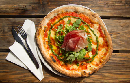 Top view of a ham and cheese pizza with rucola and pesto on a rustic wooden table