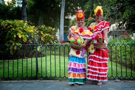 HAVANA-CUBA- DEC 4, 2018: Two Cuban woman wearing colorful cultural clothes and holding bowl of fruits and open fansの写真素材
