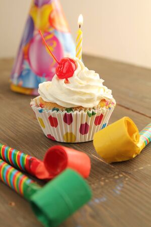 Vanilla cupcake with white frosting, cherry on top and a candle with party hat and blowers for birthday party の写真素材