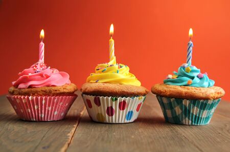Vanilla cupcake in a row with yellow, pink and blue frosting and a birthday candle on a wooden table with a orange backgroundの写真素材