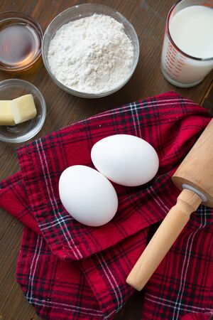 Ingredient for making delicious pancake ( eggs, flour, butter, milk with fresh maple syrup) on a red tablecloth and a rolling pinの写真素材
