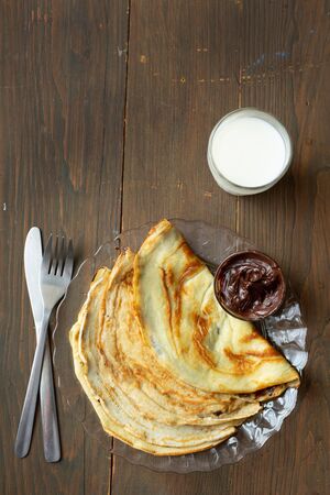 Top view of three pancakes fill with chocolate, a glass of milk and tools for breakfast on a wooden backgroundの写真素材