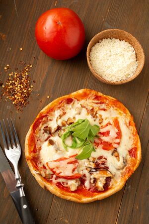 Homemade pizza with tomato, mushrooms, peppers and fresh herbs basil and arugula presented on a wooden table with cutlery and pepperの写真素材