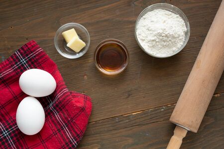 Ingredient for making delicious pancake, pie or cake ( eggs, flour, butter, milk with fresh maple syrup) on a red tablecloth and a rolling pinの写真素材