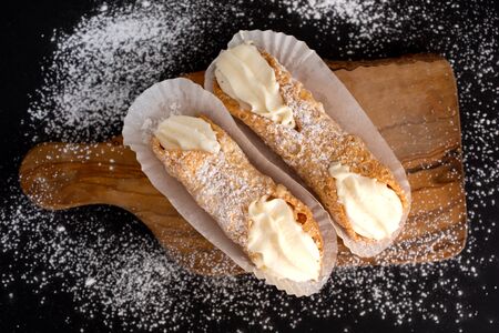 Top view of two fresh cannoli in paper on a cupboard on a black background powdered with sugarの写真素材