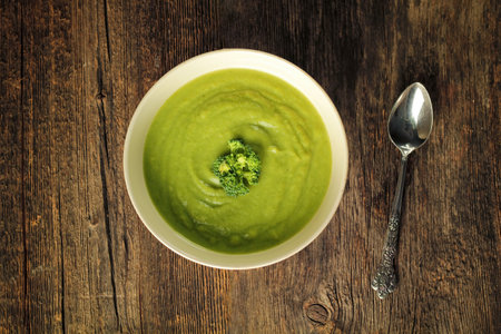 Top view of a broccoli soup in a white bowl with a spoon on a rustic wooden backgroundの写真素材