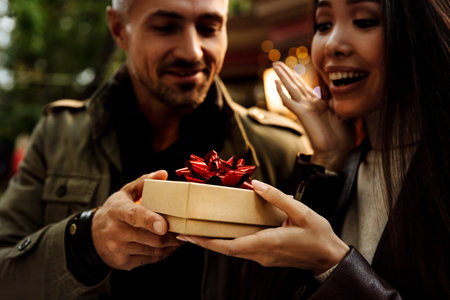 Selective focus. Couple. Holiday. Surprise. Man is giving a gift box to his woman, both are smiling;...