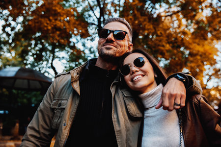 Autumn walk. Couple. Love. Man and woman in warm casual clothes and sun glasses are hugging and smiling while walking down in the parkの写真素材